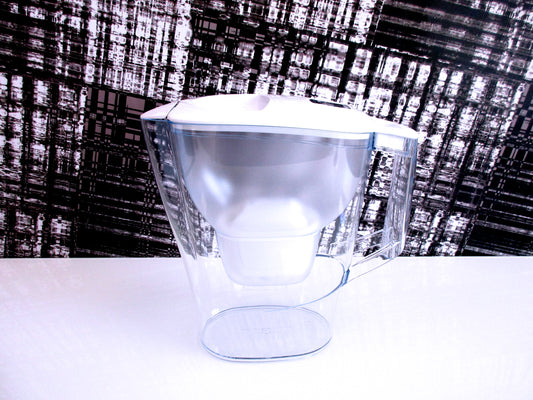A clear plastic water filter jug with a white lid, shown on a white surface against a black and white textured background.