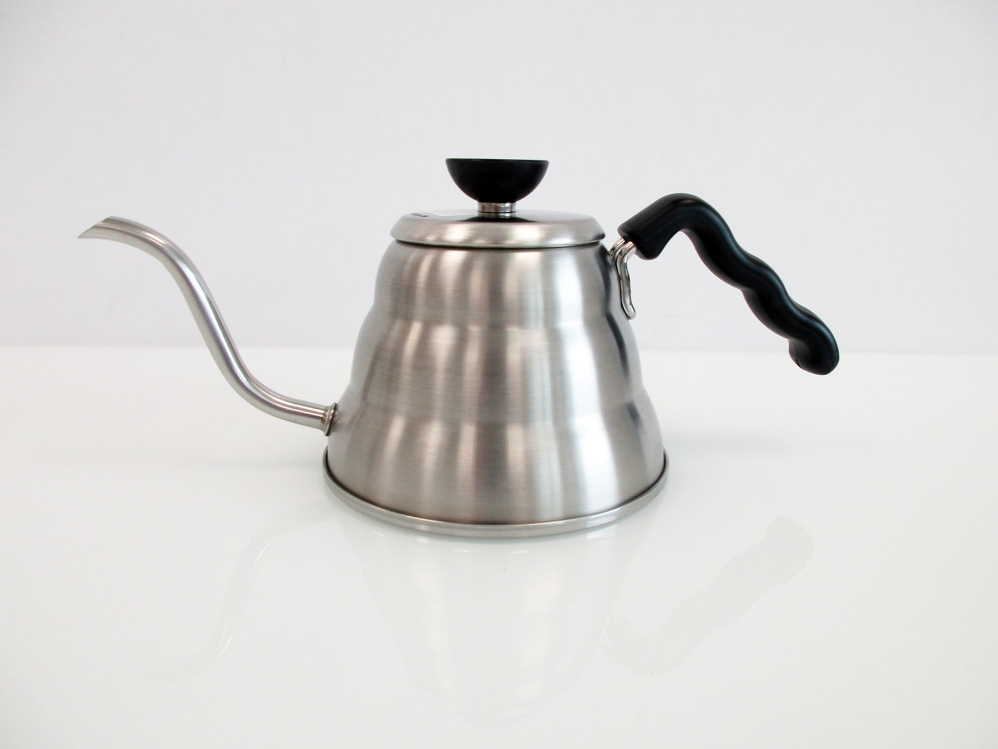 A stainless steel gooseneck kettle with a black handle and lid knob, shown on a clean white reflective surface against a white background.