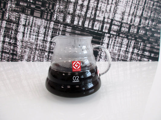 A clear glass pour-over coffee server containing hot coffee, featuring a red logo and white '02' marking, set on a white surface against a black-and-white patterned backdrop.