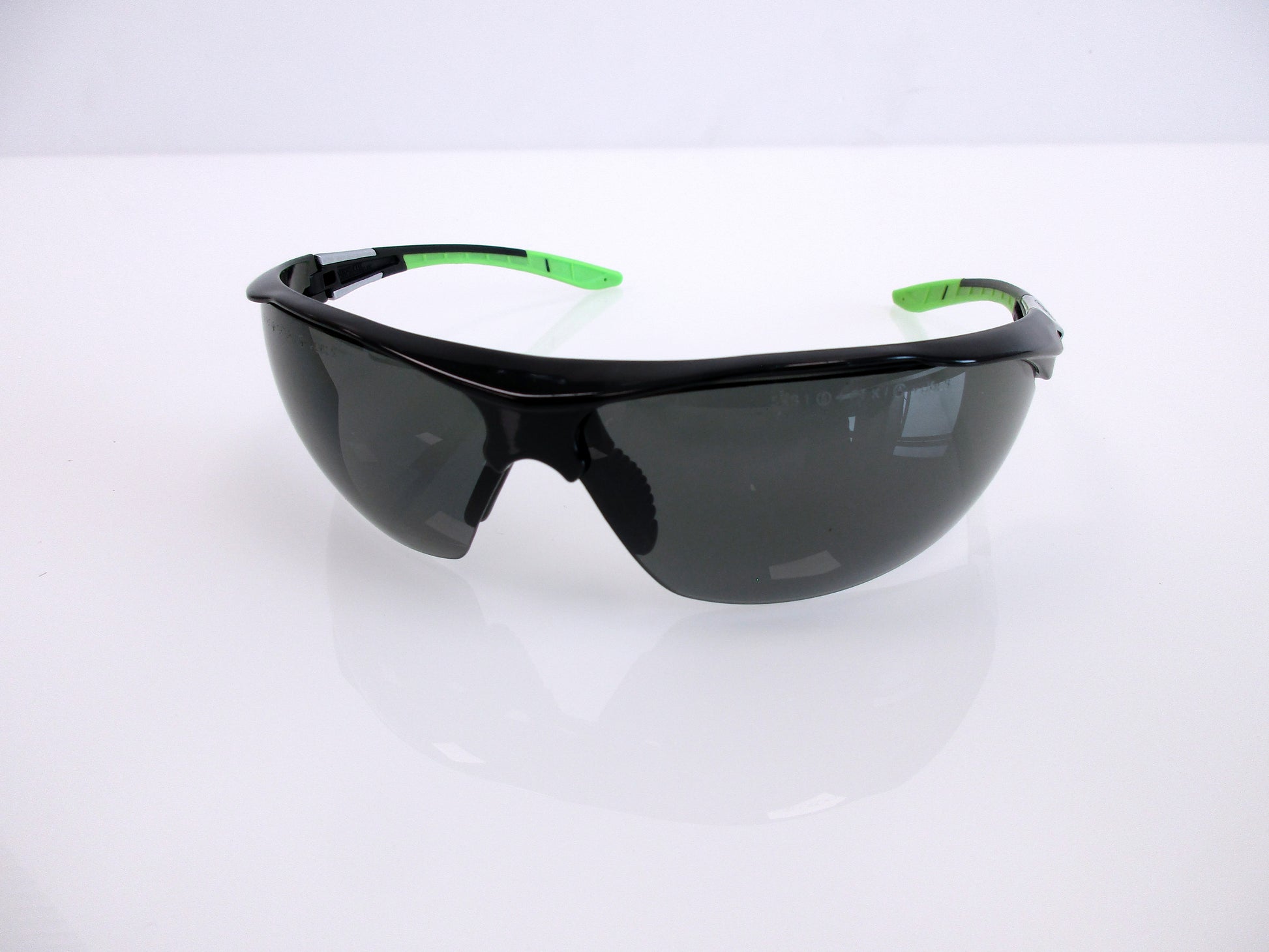 Black shield-style safety glasses with dark lenses and bright green temple tips, photographed on a clean white studio surface.