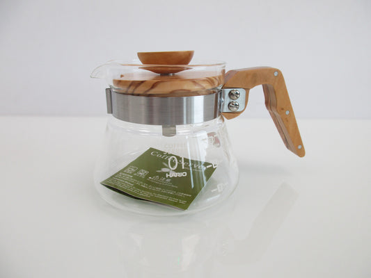 A clear glass coffee pot with a light brown wooden handle and lid, a grey metal collar, and a green paper insert inside, set against a plain white background.
