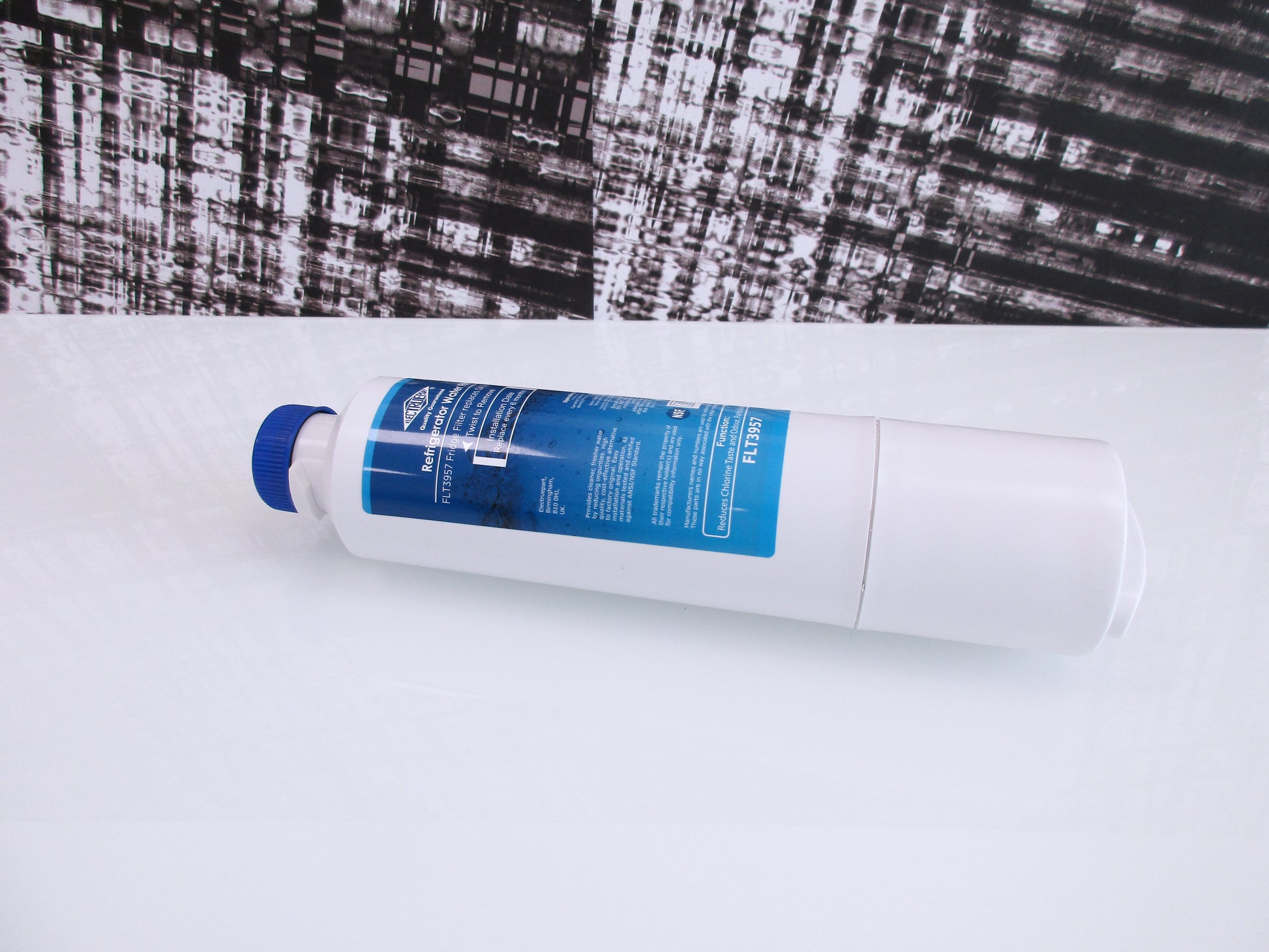 A white cylindrical water filter cartridge with a blue cap and a blue-and-white label, displaying FTL3957s, lying diagonally on a white surface with a black-and-white textured background.