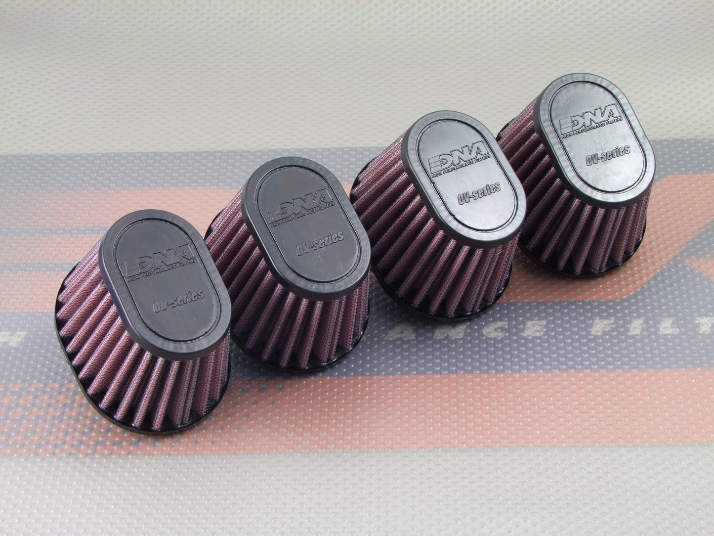 Four red pleated cone air filters with black tops, arranged in a row on a patterned surface. The brand name 'DNA High Performance Filters' is embossed on the black tops.