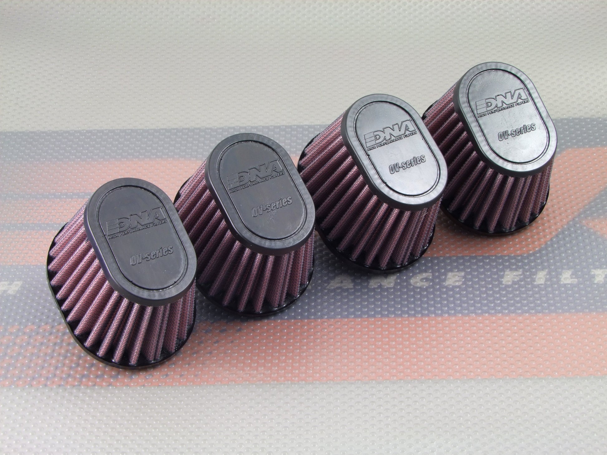 Four red pleated cone air filters with black tops, arranged in a row on a patterned surface. The brand name 'DNA High Performance Filters' is embossed on the black tops.