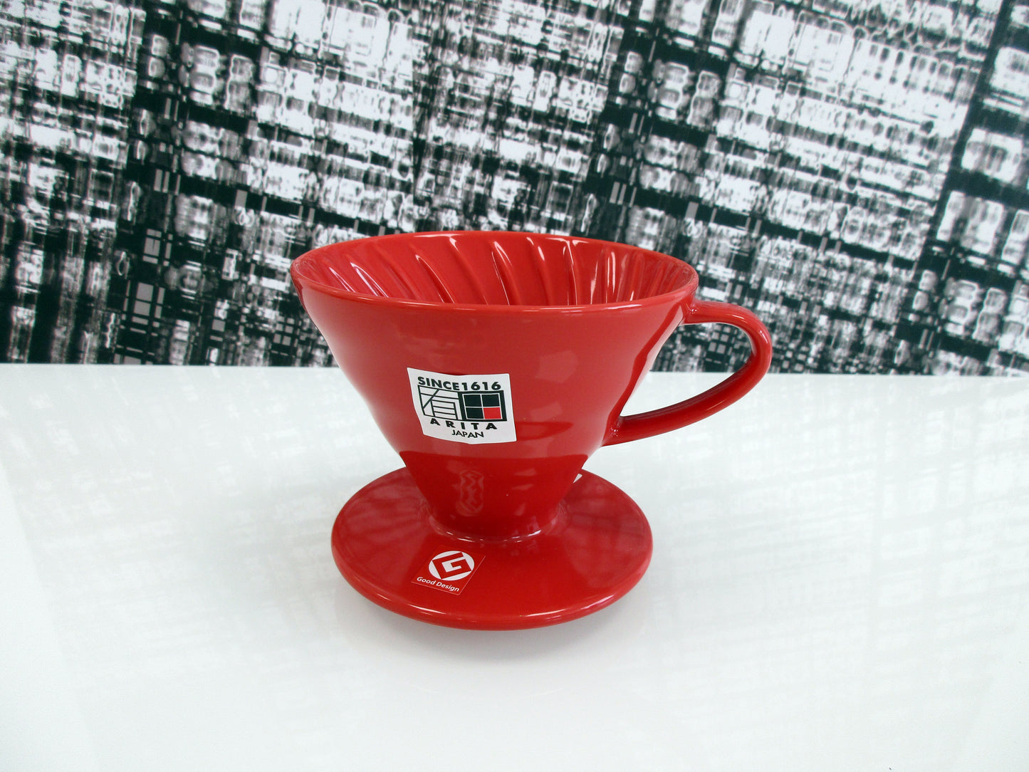 A red ceramic pour-over coffee dripper, featuring a white 'Since 1916 Arita Japan' sticker on the front, set on a white surface with a black and white textured background.