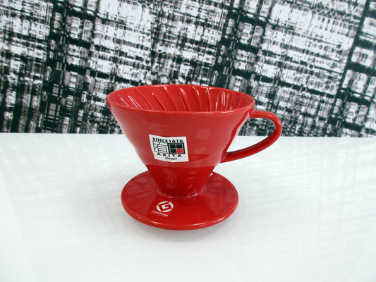 A red ceramic pour-over coffee dripper, featuring a white 'Since 1916 Arita Japan' sticker on the front, set on a white surface with a black and white textured background.