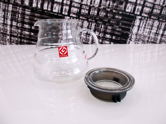 A clear glass coffee server with a small red logo sticker on the front, shown next to a dark grey silicone lid, on a white reflective surface.