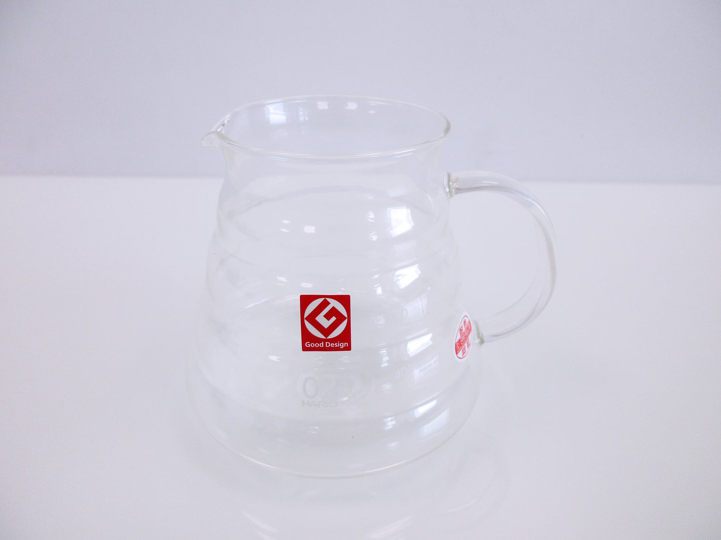 A clear glass coffee pitcher with a handle and spout, featuring a red square logo on the front, set against a plain white background.