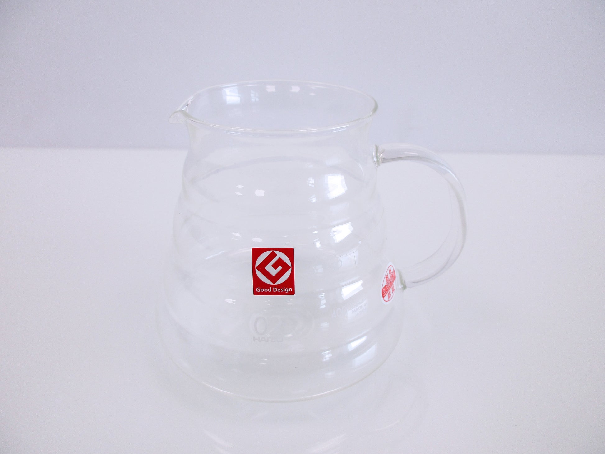 A clear glass coffee pitcher with a handle and spout, featuring a red square logo on the front, set against a plain white background.
