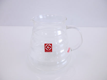 A clear glass coffee pitcher with a handle and spout, featuring a red square logo on the front, set against a plain white background.