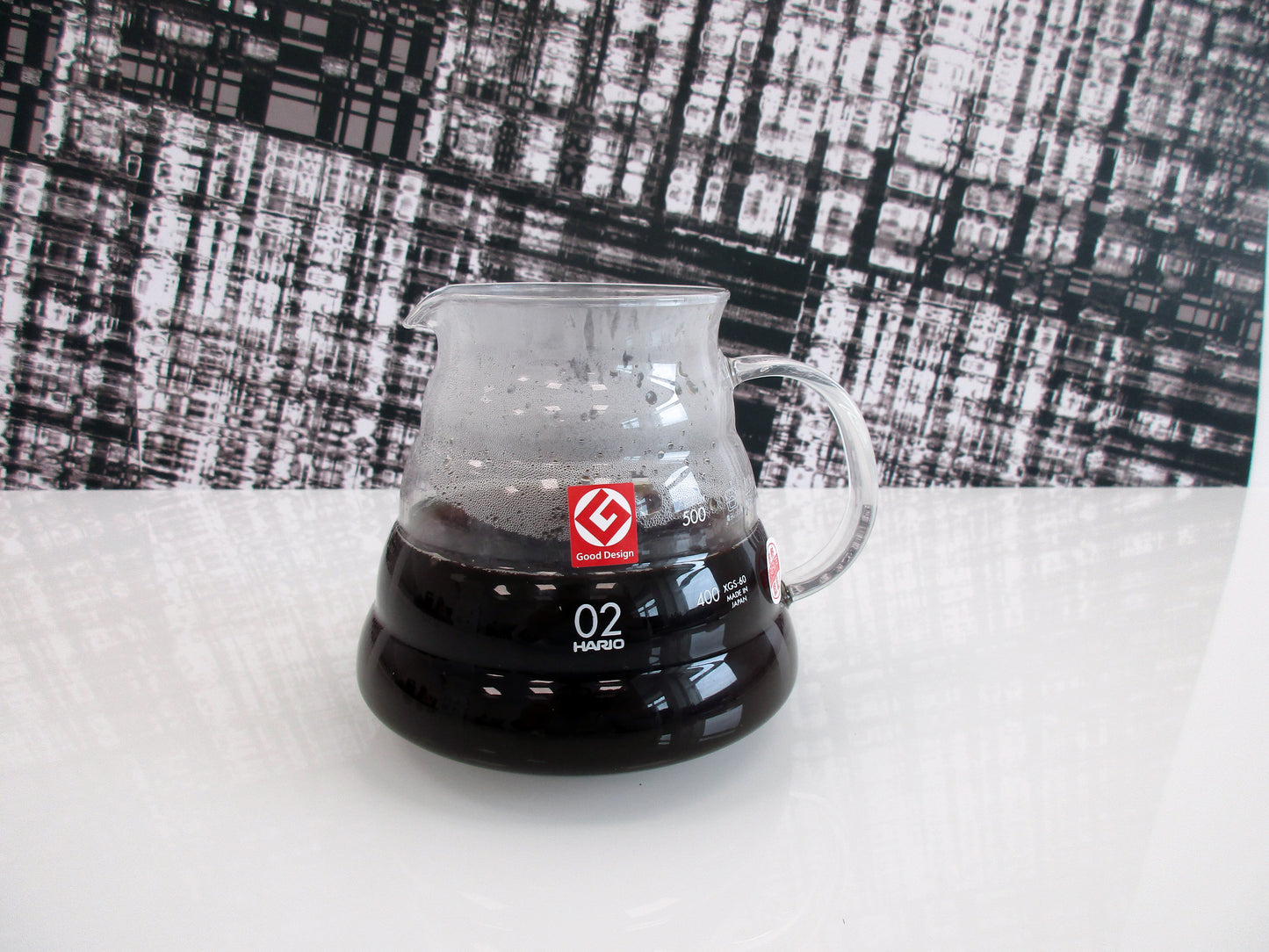 A clear glass pour-over coffee server containing hot coffee, featuring a red logo and white '02' marking, set on a white surface against a black-and-white patterned backdrop.