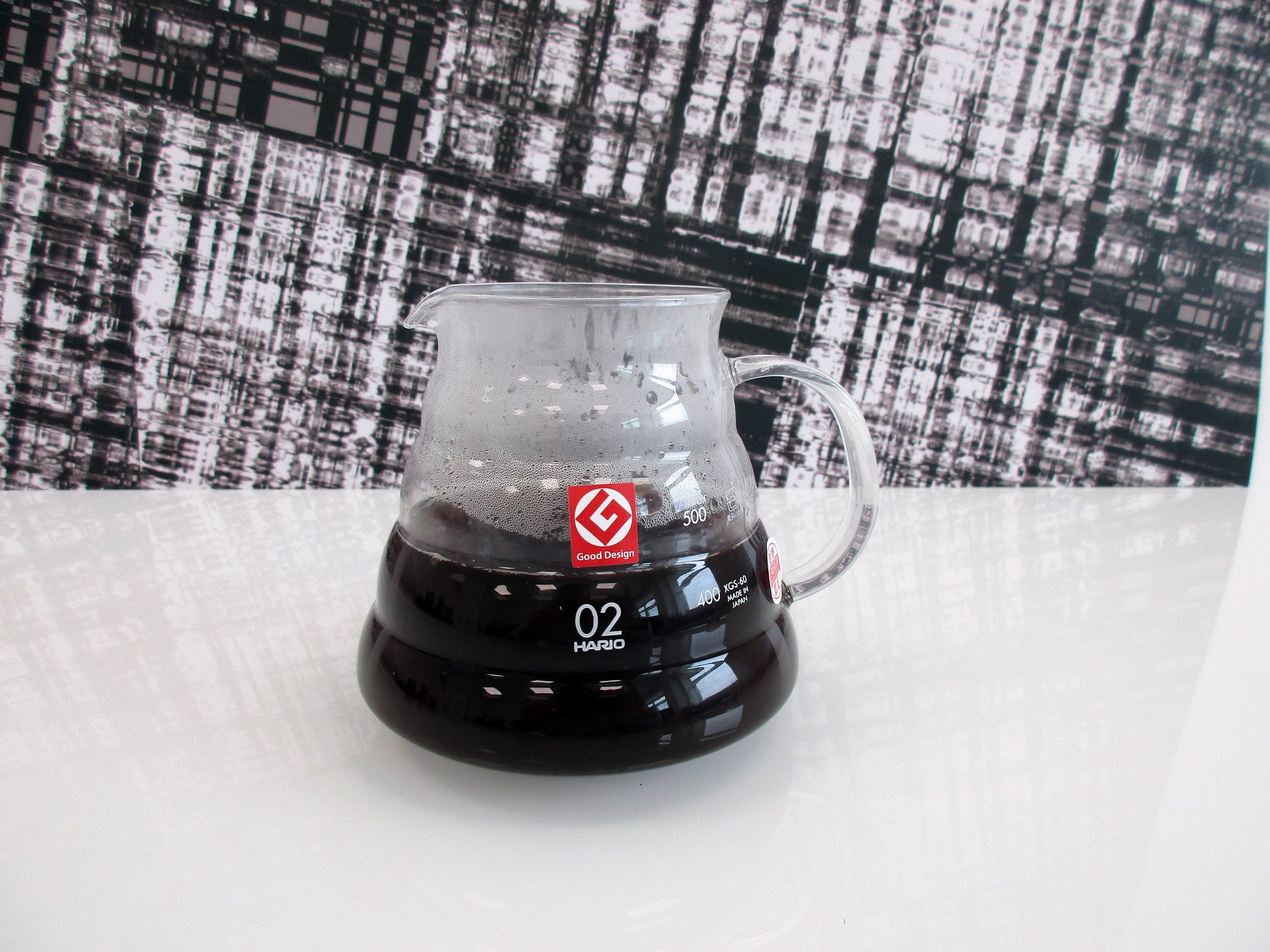 A clear glass pour-over coffee server containing hot coffee, featuring a red logo and white '02' marking, set on a white surface against a black-and-white patterned backdrop.
