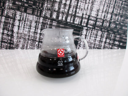 A clear glass pour-over coffee server containing hot coffee, featuring a red logo and white '02' marking, set on a white surface against a black-and-white patterned backdrop.